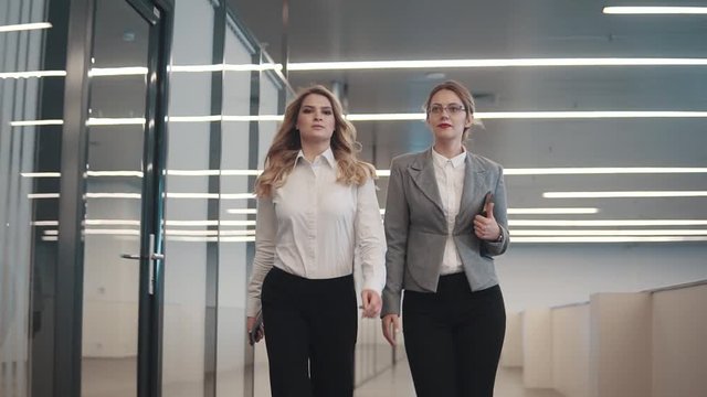 two young business women walking along the corridor of an office building. girls in business clothes in the workplace. slow motion