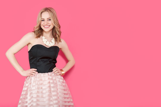 Attractive blonde young woman in elegant party dress and golden jewelry. Girl posing on a pastel pink background. Fashion photo.