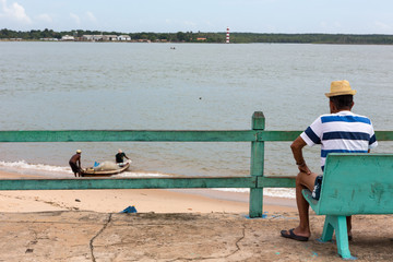 One man sitting on bench watches two fishermen fishing on the Amazon river in the bay of the Marajó island.