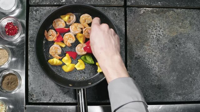 Top view of unrecognizable cook adding fresh chopped bell peppers into pan with shrimps frying in oil