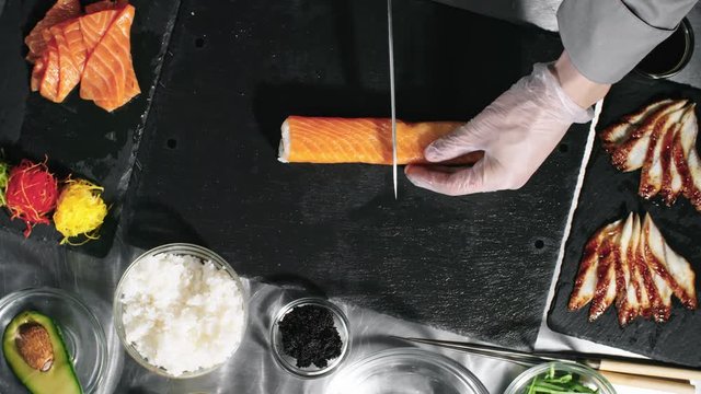 Top view of unrecognizable cook in gloves wetting knife in water and cutting sushi rolls