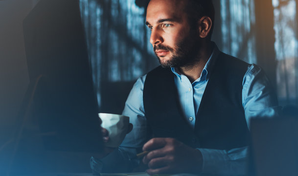 Bearded young businessman working on modern office. Consultant man thinking looking in monitor computer. Manager typing on keyboard in coworking workplace, startup project concept in night with coffee