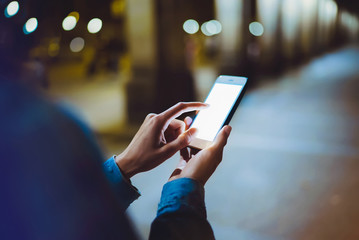 Woman pointing finger on blank screen smartphone on background bokeh light in night atmospheric city, hipster blogger using in hands gadget mobile phone, mockup street, online wifi internet concept