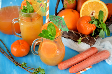 Orange and carrot juice in jugs decorated with mint, cocktail straw and umbrella on blue background