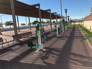 Long shot of an outdoor gym, facing the sea, on a sunny day. In the distance people walking, running, walking, cycling,