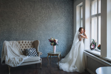 A beautiful bride in a lace dress stands near a window in a gray studio. Portrait of a blonde bride.