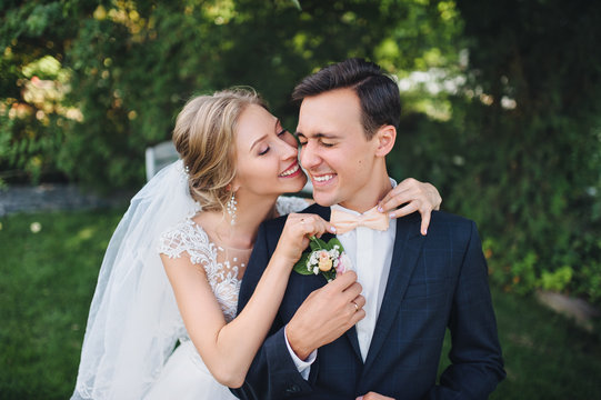 A beautiful bride in a lace dress adjusts a bow tie to a stylish groom. Emotional wedding in a green garden.