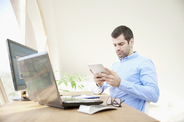 Professional man with digital tablet, laptop and personal computer. Portrait of young businessman looking thoughfully while sitting at office desk and using digital tablet. 