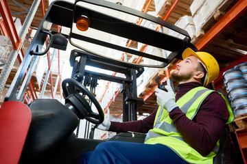 Side view portrait of young man sitting in forklift and using walkie-talkie while moving goods in...