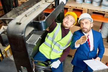 High angle portrait of warehouse manager holding clipboard talking to worker wearing hardhat and...