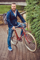 A bearded male with a stylish haircut dressed in casual clothes with a backpack, sitting on a retro bicycle in a city park.