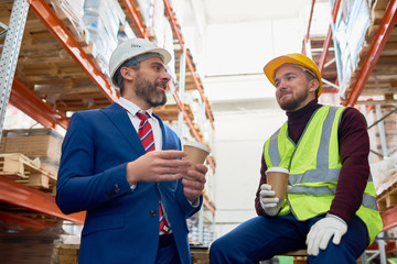 Waist up portrait of two warehouse workers taking break sitting on pellets drinking coffee and...