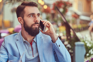Close-up portrait of a fashionable bearded businessman with a stylish haircut, speaking by phone, drinks a glass of a juice, sitting in a cafe outdoors.