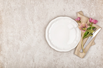 Empty white plate, cutlery set with vintage style linen napkin and purple flowers on stone background. Top view, flat lay and copy space
