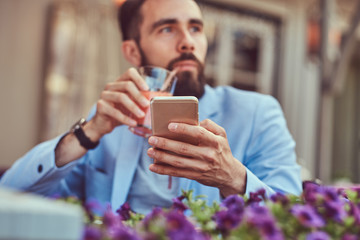 Portrait of a fashionable bearded businessman with a stylish haircut, drinks a glass of a juice and holds a smartphone, sitting in a cafe outdoors.