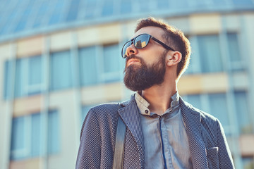 Close-up portrait of a bearded male wearing casual clothes and sunglasses, standing in a city against a skyscraper.