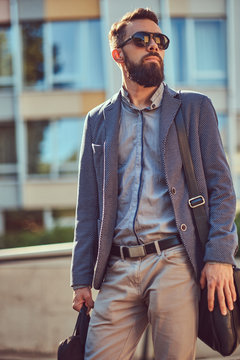 Portrait of a bearded worker wearing casual clothes and sunglasses, with a bag, standing in a city street against a skyscraper.