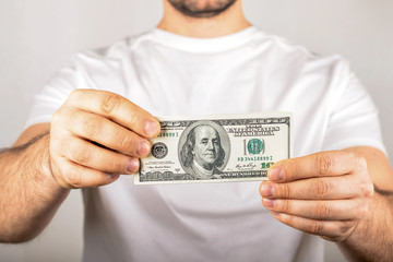 a man in a white T-shirt with a dark stubble holds with both hands and holds out a hundred dollar...