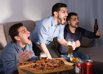Exalted males watching tv at home, enjoying beer and pizza