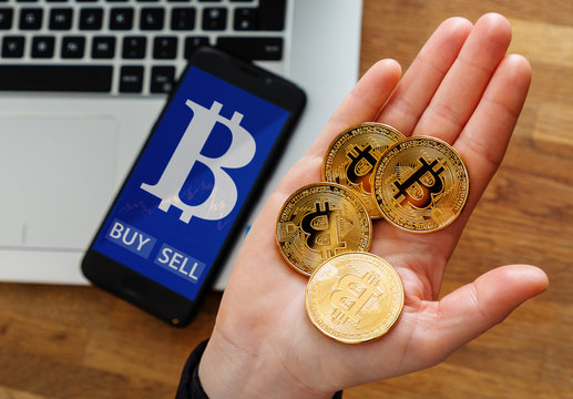 human's hand holding gold coin of Bitcoin and Virtual currency symbol on mobile app screen with big BUY and SELL buttons.
