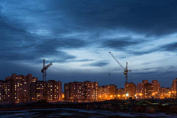 building site with two tower cranes against the background of multi-storey houses and a blue sky, night scene