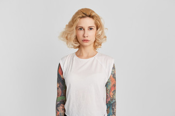 Indoor shot of young blonde female wears t-shirt with tattoos over her arms looks directly into...