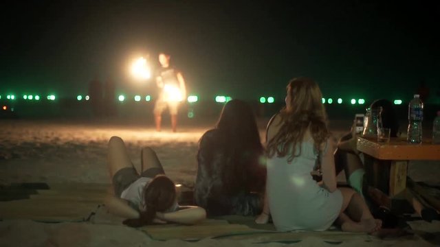 Fire artist performs on a beach  bar young women  watching his performance on a summer night with their face to the water drinking alcohol dark skies Green light in the background