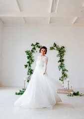 Fototapeta premium The young bride in an elegant wedding dress stands on a white background decorated with green foliage