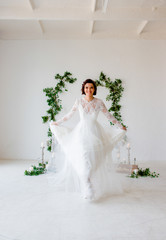 Fototapeta premium A young bride whirl in a white dress on a white background decorated with green leaves