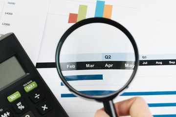 Business performance review by month or quarter concept, hand holding magnifying glass on Q1 and Q2 report with calculator and bar graph on table
