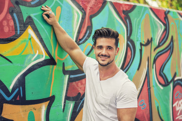 Fototapeta na wymiar Attractive muscle man leaning on colorful graffiti wall, wearing white t-shirt, smiling to camera