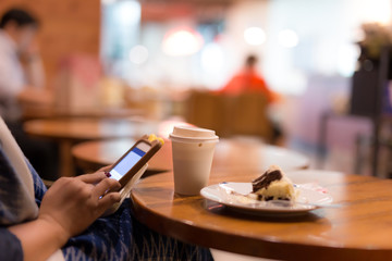 Selected focus cup of coffee with woman using cell hone and eating cake on wooden table