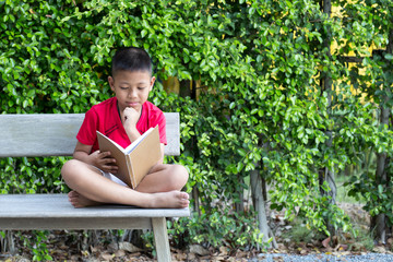 Little boy sitting on wooden bench hand on his chin reading a book