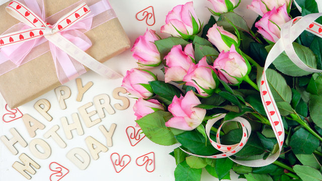Mother's Day overhead with gift and pink roses on white wood table background with wooden letters spellling Happy Mother's Day.