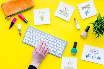 Beauty blogging concept. Work desk with keyboard, cosmetics and social media icons on yellow desk top view. Hand is typing