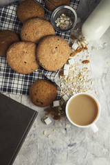 Сup of coffee with milk and oatmeal cookies on a stone background. White cup, book, oatmeal, raisins, sugar cubes, bottle of milk. Scene from breakfast