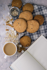 Сup of coffee with milk and oatmeal cookies on a stone background. White cup, notebook, oatmeal, raisins, sugar cubes, bottle of milk. Scene from breakfast