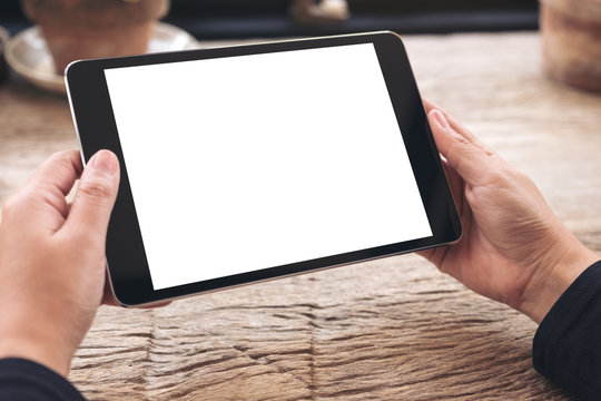 Mockup image of hands holding black tablet pc with blank white screen on vintage wooden table