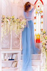 Young dreaming girl in dress with a train blue color sitting on flowers and window background of the fairytale interior 