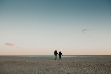 Man and woman walking by the beach sand at sunset, holding hands, with the sea and a blue sky in the background.
