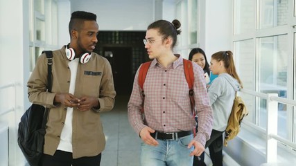 Caucasian hipster and african-american students are going down in college big glassy corridor discussing something waving hands to female classmates and move on