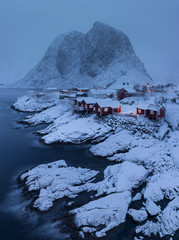 fishing village on the shore of fjord with typical norwegian red cabins covered by snow and mountain in background, hamnoy, lofoten, norway