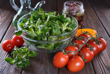 Leaves of corn salad in a glass bowl, a cherry tomato branch, pepper, olives in a jar and olive oil in an oil can ready for salad cooking are on a wooden table