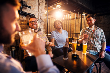 Four happy casual multicultural friends having fun and drinking beer while sitting in the pub.