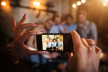 Close up focus view of woman hands and mobile while taking a photo of four happy friends relaxing in the pub.