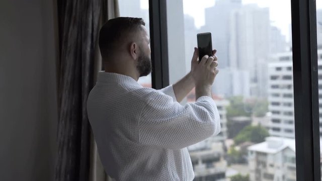 Man standing in his hotel room and captures view from window on smartphone
