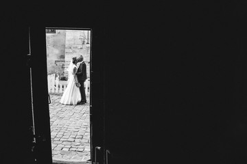 Beautiful wedding couple is hugging in the old city. White cafe fence and cobblestone road on background. Bride in satin lace dress and happy groom. Ancient street photo taken from the door frame.
