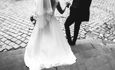 Wedding blonde couple is walking in old city. Bride in lace satin dress and groom are holding hands...
