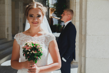 Wedding blonde couple is walking in the old city. Groom is standing near the ancient stone walls columns. Bride in lace satin dress is holding pink rose bouquet. Sunny love story in the medieval town.
