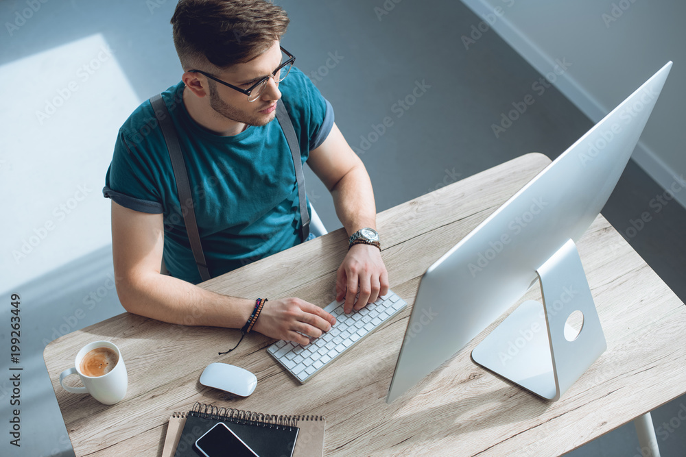 Poster overhead view of young man in eyeglasses typing on keyboard and working with desktop computer